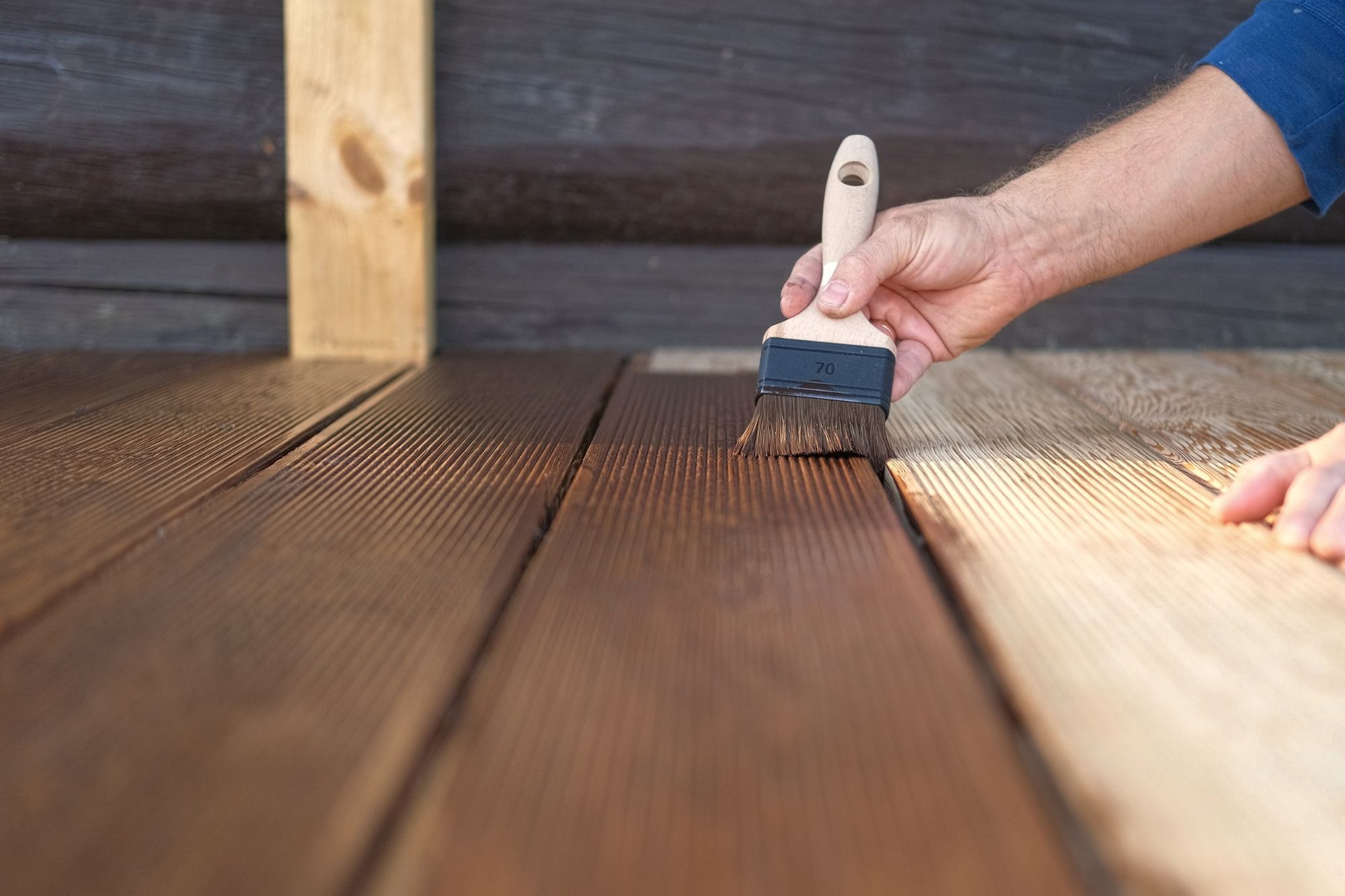 Person’s hand with a paintbrush staining wooden deck planks outdoors.