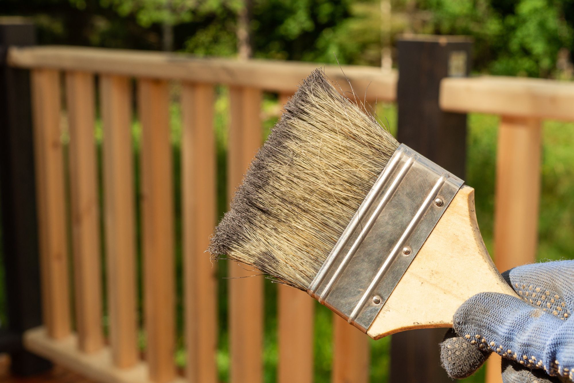 Close-up of a paintbrush with dry bristles and metal ferrule, outdoors beside a wooden railing.